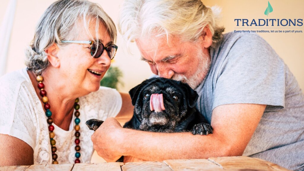 An elderly couple sitting next to each other outside, both looking down at their black pug. The man has his hand around the dog and is kissing the top of the dog's head.