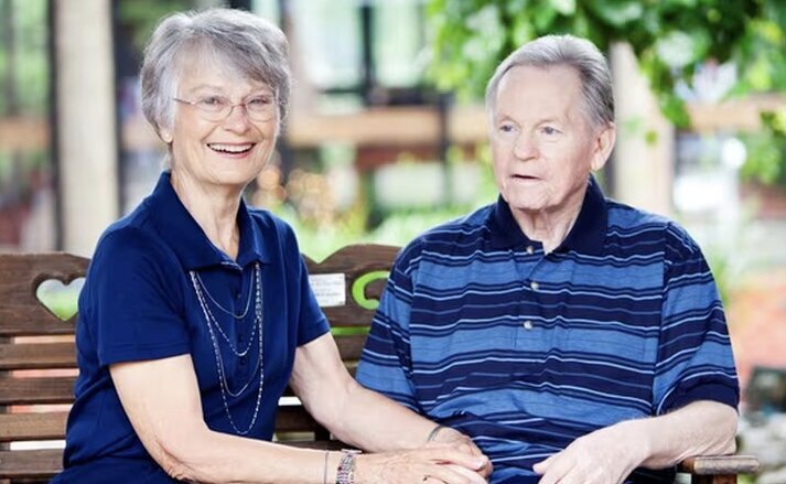 A happy couple sitting on a bench outside enjoying the sunshine. Traditions at Brookside.