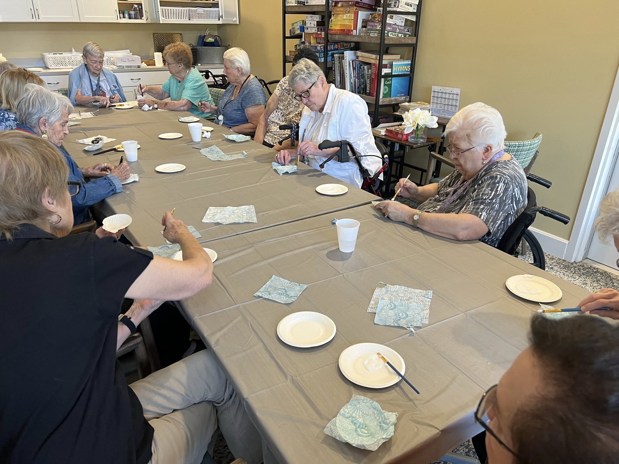 A group of seniors is sitting around a gray table, working on an art project with white plates and green patterned napkins.