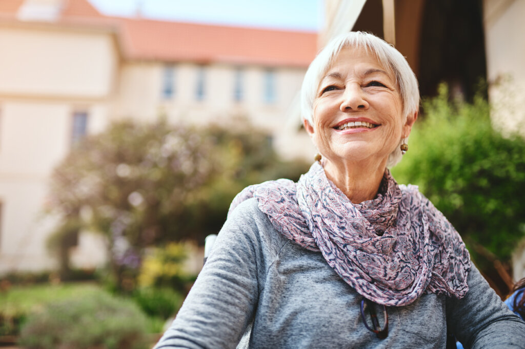 A senior woman relaxing wearing a gray long sleeve and pink scarf, outside of a senior living community.