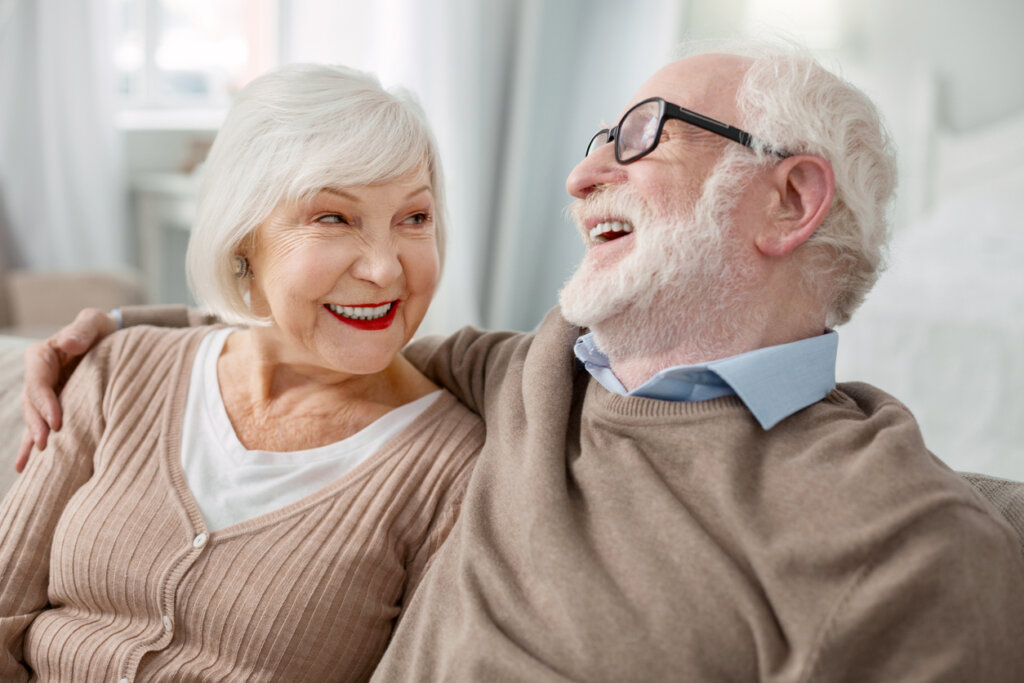 An elderly couple sitting together on a couch, the elderly man has his hand over his wife's shoulder.