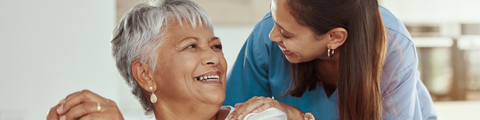 An older woman with grey hair is seated, looking upward and smiling. Leaning over her is a woman wearing blue scrubs, smiling