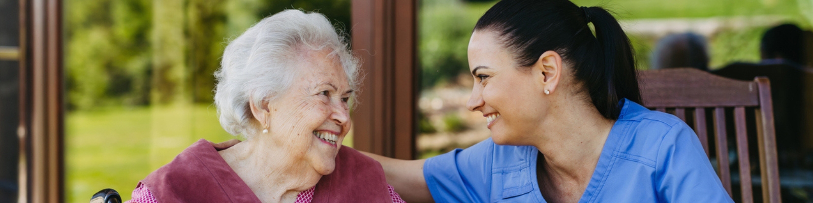 n older woman with short white hair & a caregiver wearing blue scrubs, looking at each other & smiling outside on a patio